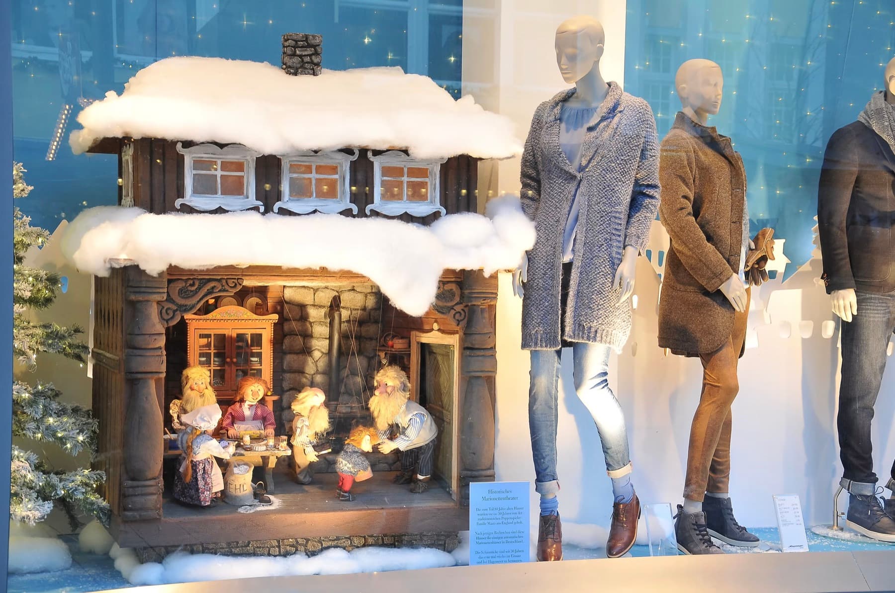 Beleuchtetes Marionettenhaus mit Puppenszene im weihnachtlichen Schaufenster neben Schaufensterpuppen